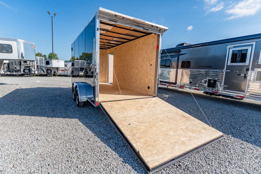 Open cargo trailer with its wooden interior exposed and the ramp lowered for loading, parked on a gravel lot with other trailers nearby.