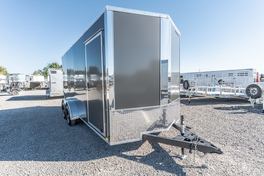 Silver enclosed cargo trailer with diamond-plate front on a gravel lot, hitch attached, and other trailers in the background.