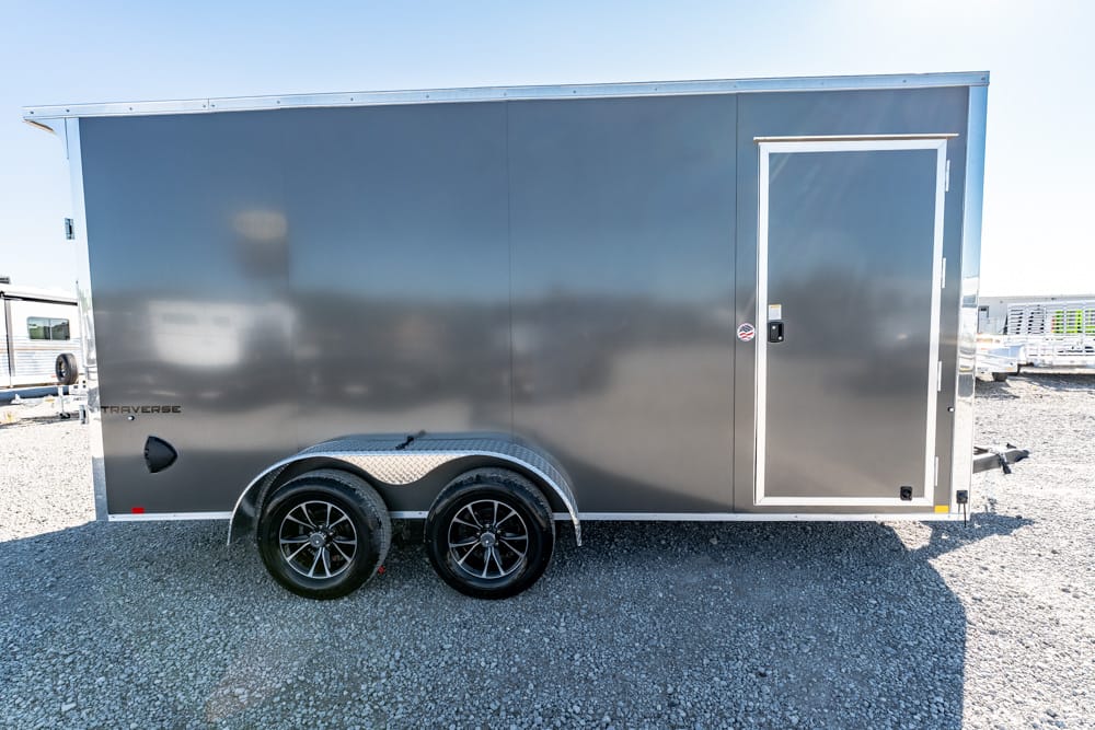 Gray enclosed cargo trailer with tandem wheels and a side entry door on a gravel lot.