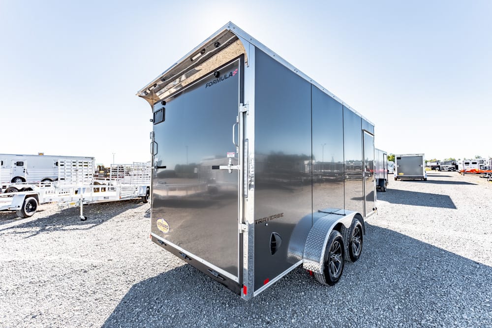 Gray enclosed cargo trailer with tandem wheels parked on a gravel lot, sunny day