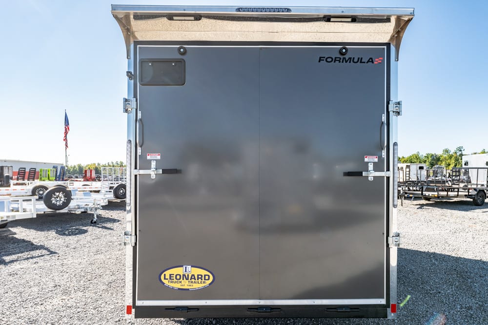 Rear view of a dark gray Formula trailer with Leonard Truck & Trailer logo, parked in a gravel lot with other trailers and a flag nearby.