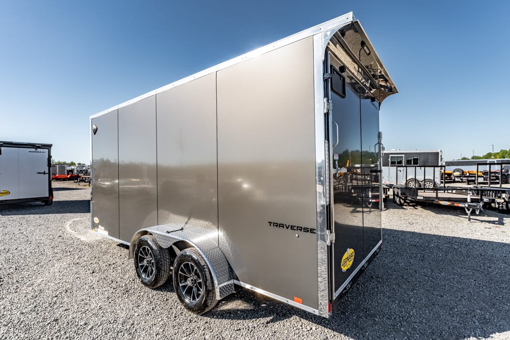 Silver enclosedTraverse trailer with dual axles and black rims parked on a gravel lot, rear ramp door closed and blue sky above.