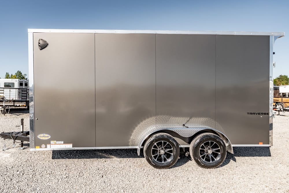 Gray enclosed cargo trailer with tandem axles and black wheels, parked on gravel.