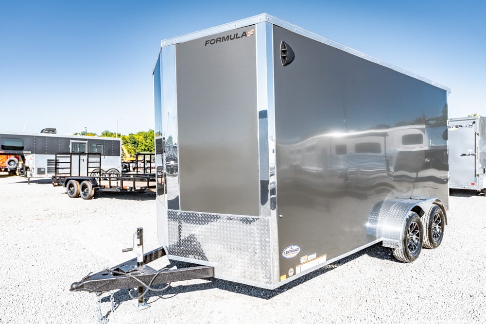 Shiny gray enclosed cargo trailer with dual axles and diamond-plate front parked on a gravel lot, hitch at the front.