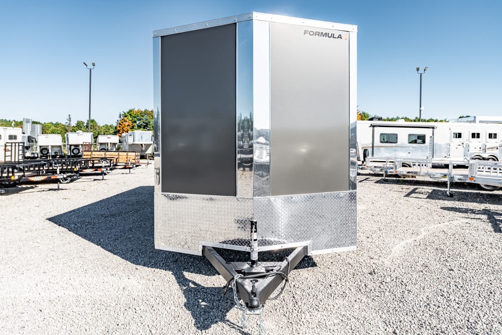 Front view of a shiny silver enclosed trailer with diamond-plate protection at the hitch, parked on a gravel lot with other trailers in the background.