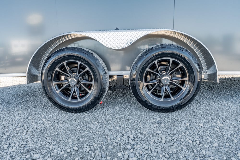 Dual-axle trailer wheels with black rims and diamond-plate fenders on a gravel lot, under a metallic trailer wall.