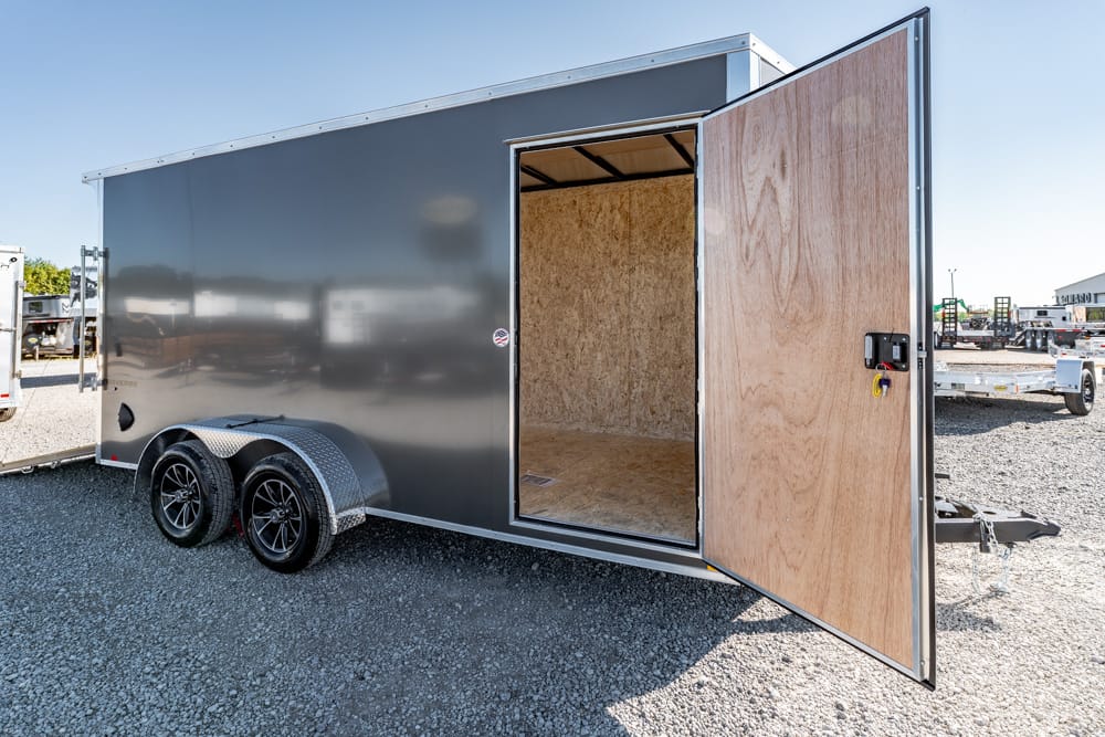 Gray enclosed cargo trailer with dual axles and an open rear door showing plywood interior on a gravel lot