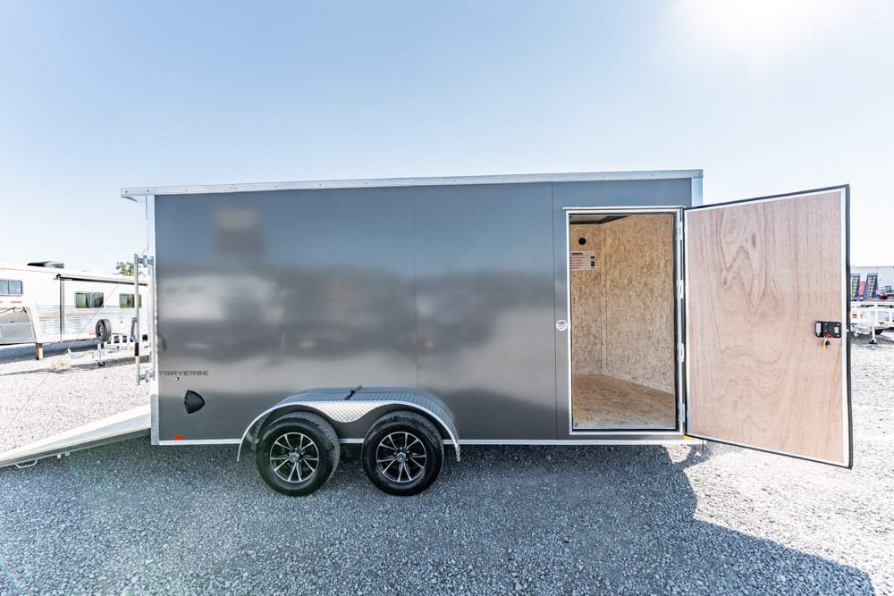 Silver enclosed cargo trailer with its side door open, revealing plywood interior on a gravel lot with dual axles and ramp nearby.