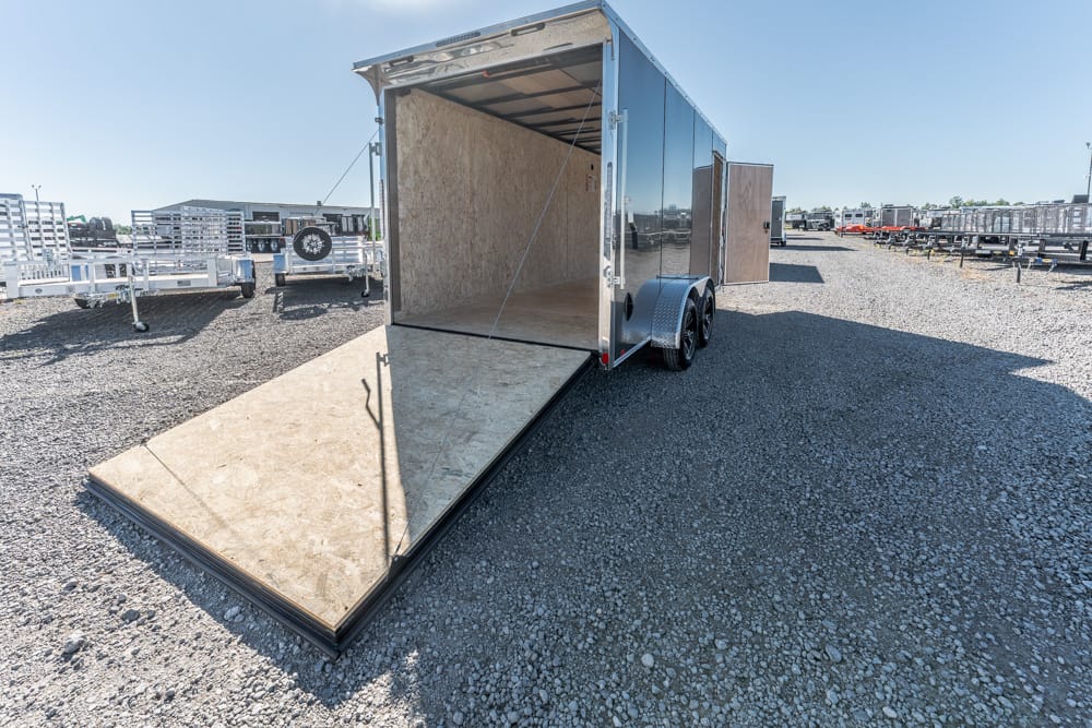 Open enclosed cargo trailer with a lowered ramp on a gravel lot, surrounded by other trailers.