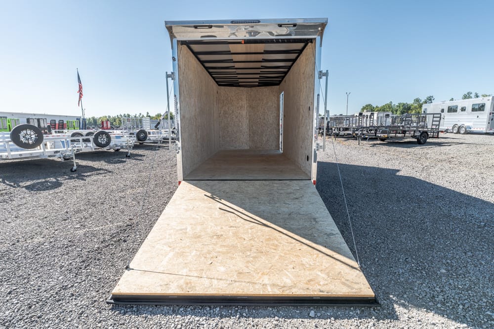 Open cargo trailer with ramp down, ready for loading in a gravel lot. The interior is unfinished wood panels, with other trailers visible in the background.