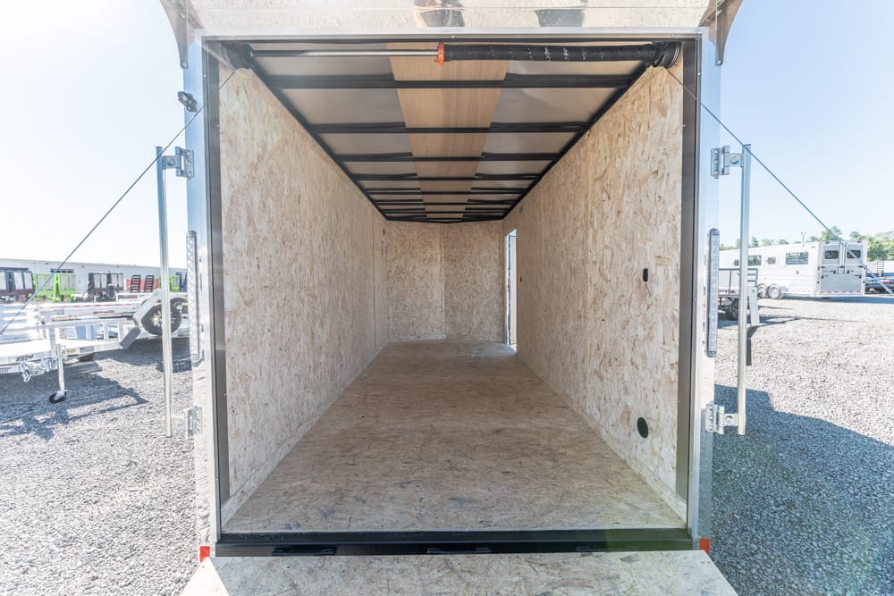 Interior of an empty enclosed trailer with plywood walls and floor, open rear doors, daylight outside on a gravel lot.
