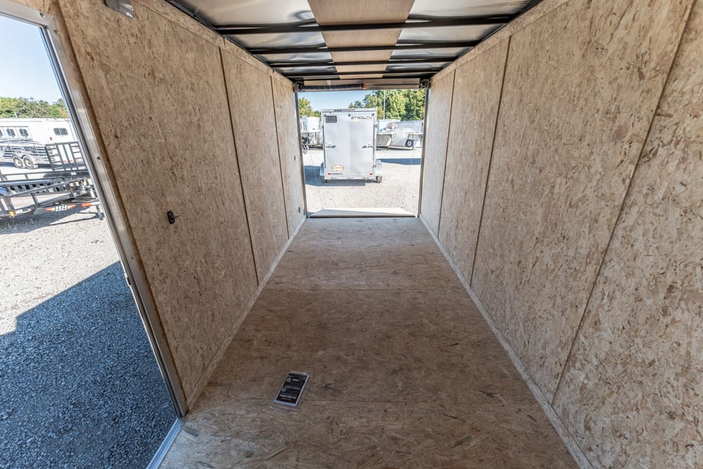 Interior of an empty plywood trailer hallway, looking toward the open rear with other trailers visible outside on a gravel lot.