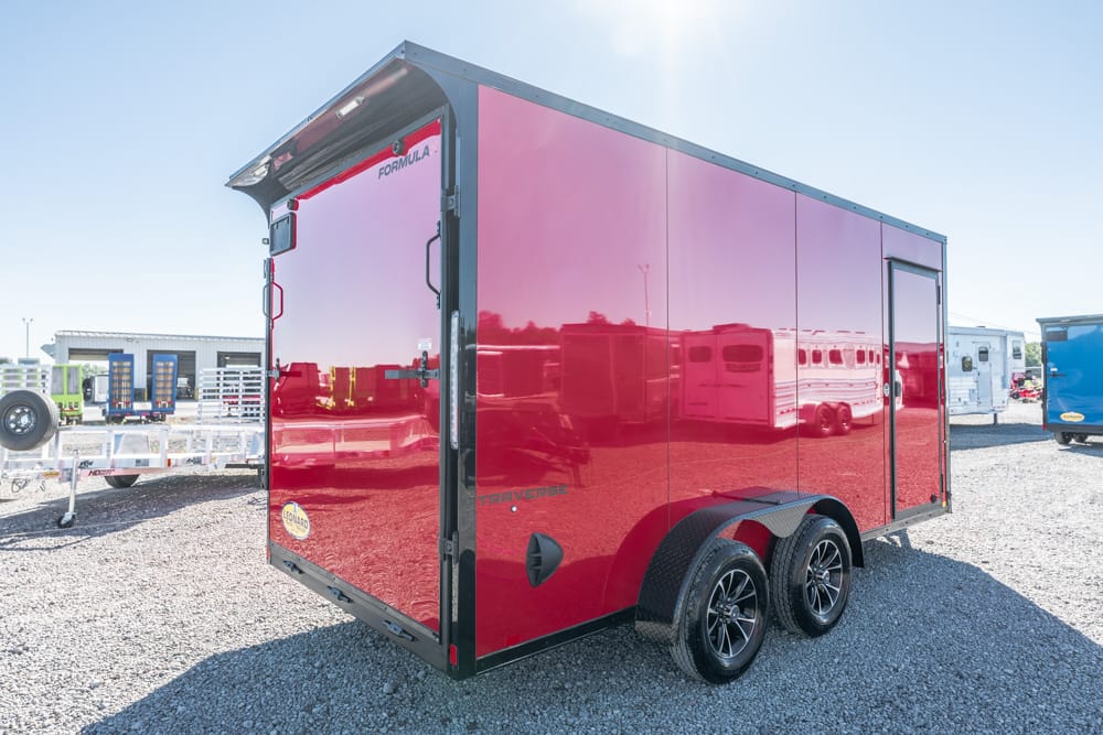 Glossy red, dual-axle enclosed trailer with black trim parked on a gravel lot, reflecting surroundings in its surface.