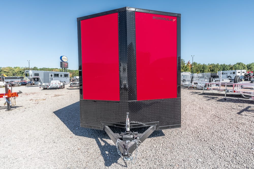 Enclosed red and black cargo trailer with a V-shaped front hitch on a gravel lot at a trailer yard.