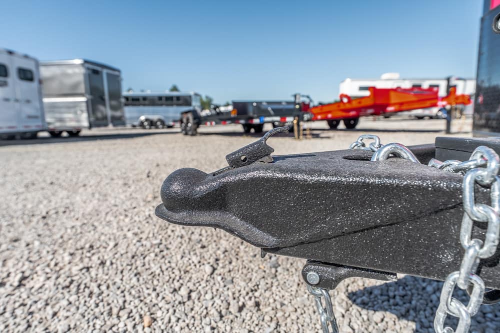 Close-up of a black trailer coupler with safety chains, in a gravel lot with trailers in the background.