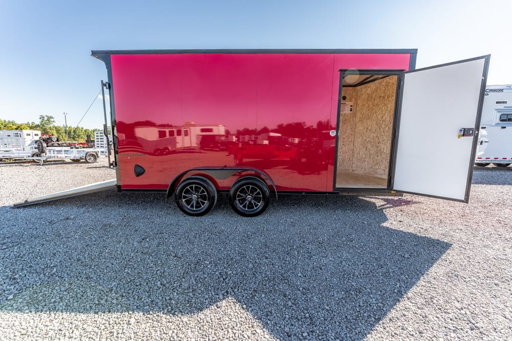Red enclosed trailer on a gravel lot with its side door open and ramp lowered, showing an empty interior.