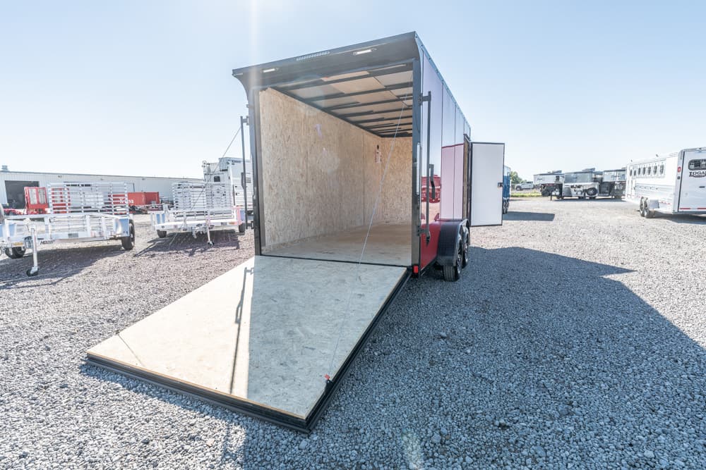 Open red cargo trailer with a lowered wooden ramp in a gravel lot, surrounded by other trailers.
