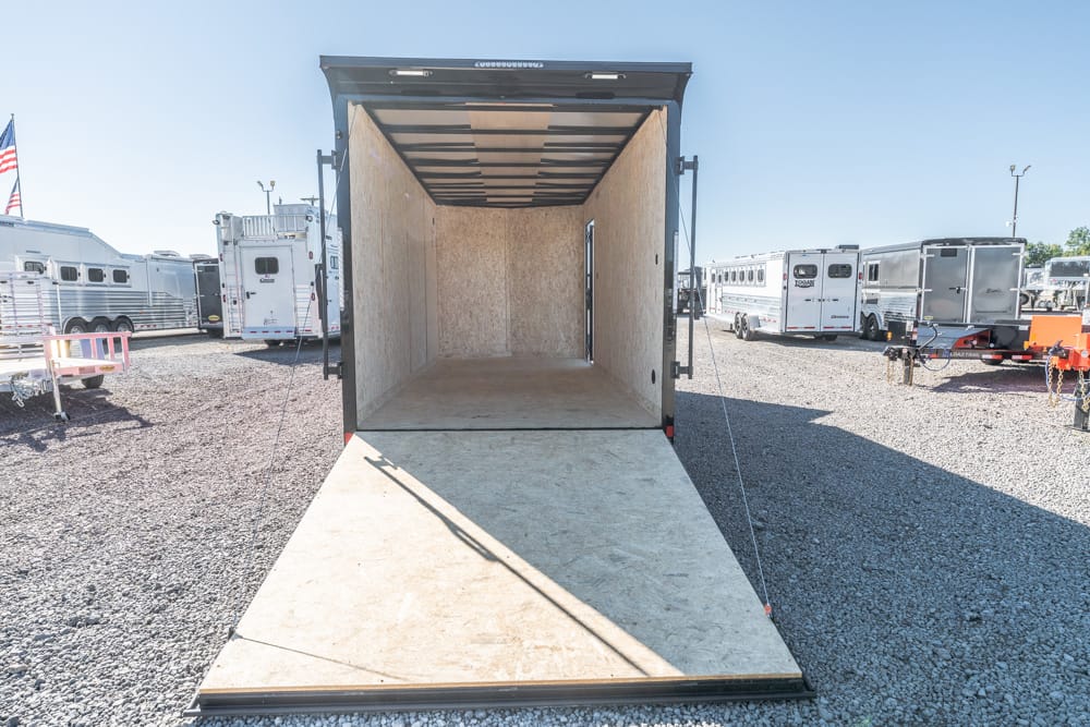 Open cargo trailer with its ramp down, empty interior visible, parked in a gravel lot among other trailers on a sunny day.
