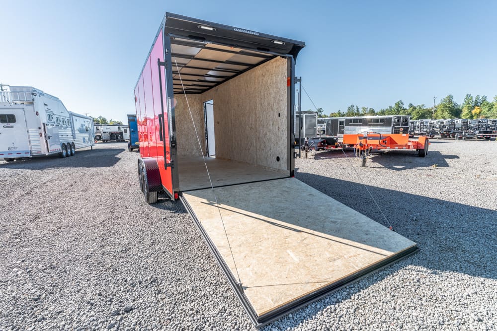 Red cargo trailer with its rear ramp down, showing an empty plywood interior on a gravel lot.