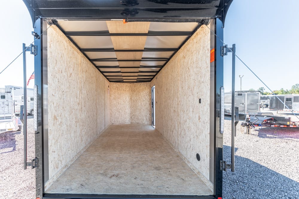 Empty enclosed trailer interior with plywood walls, viewed from rear open doors, showing a bare wooden floor and ceiling supports.