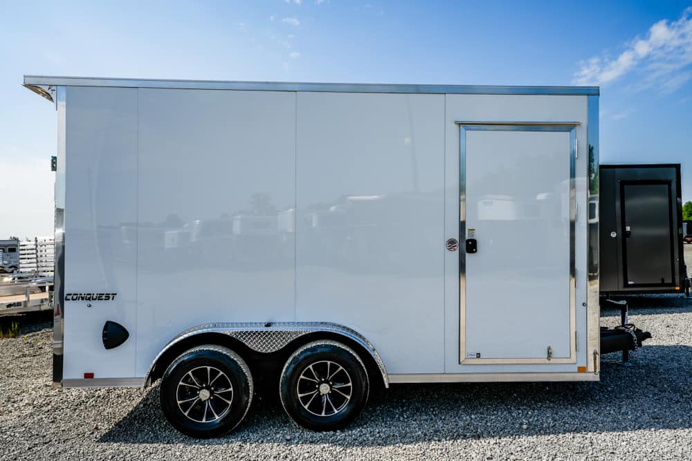 Side view of a white, tandem-axle enclosed cargo trailer with chrome diamond-plate fenders on a gravel lot; branding reads 'Conquest' near the rear.