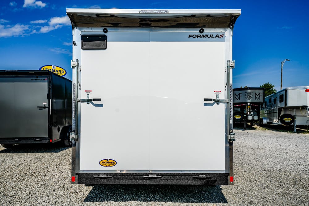 Rear view of a white enclosed cargo trailer with double doors, metal framing, and FORMULA branding, parked on a gravel lot under a blue sky.