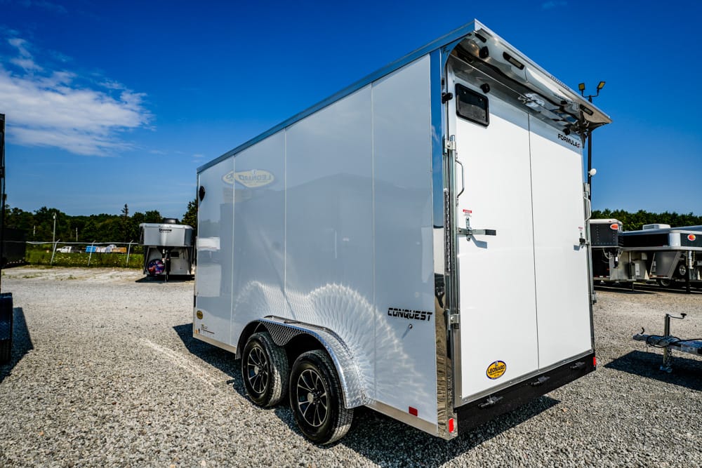 Back view of a white enclosed cargo trailer with double doors, dual axles, and diamond-plate fenders on a gravel lot under a blue sky