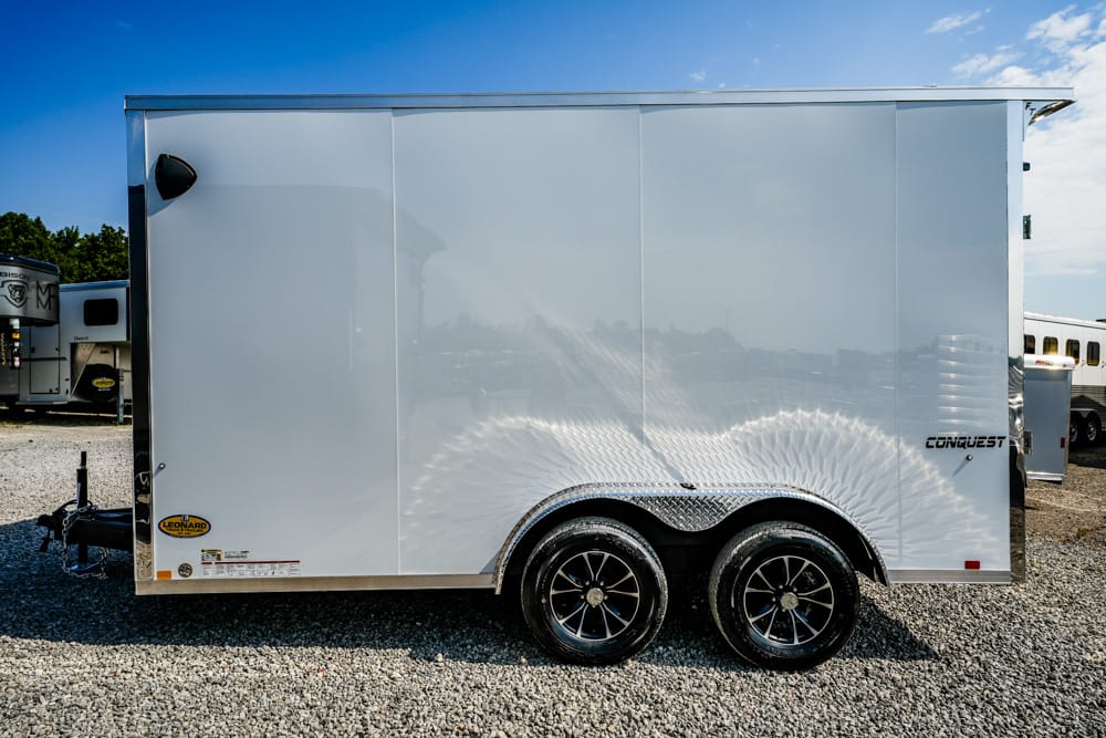 White enclosed trailer with two axles and diamond-plate fenders on gravel. A clean, gleaming cargo trailer parked on a lot.