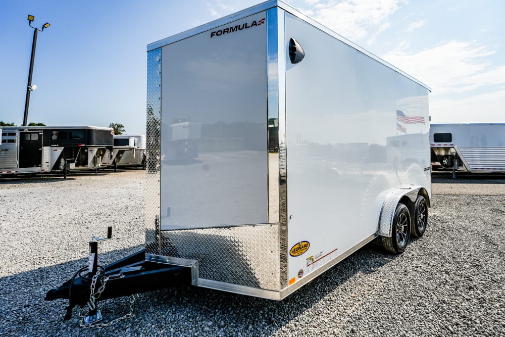 Silver enclosed cargo trailer with dual axles and a diamond-plate front on a gravel lot.