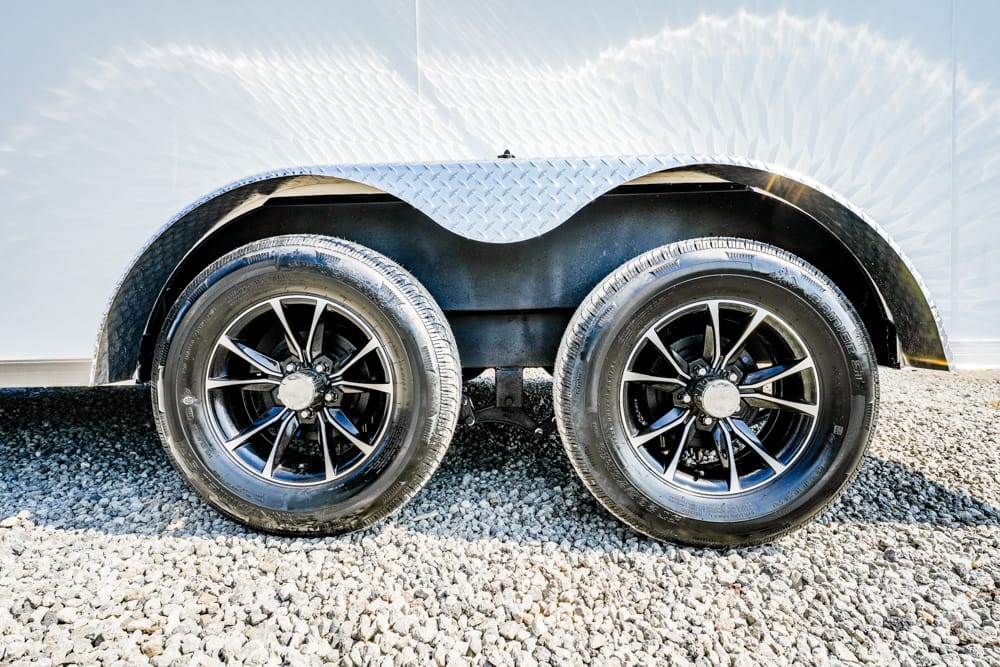 Dual-axle trailer with black alloy wheels and a diamond-plate fender on a gravel surface.