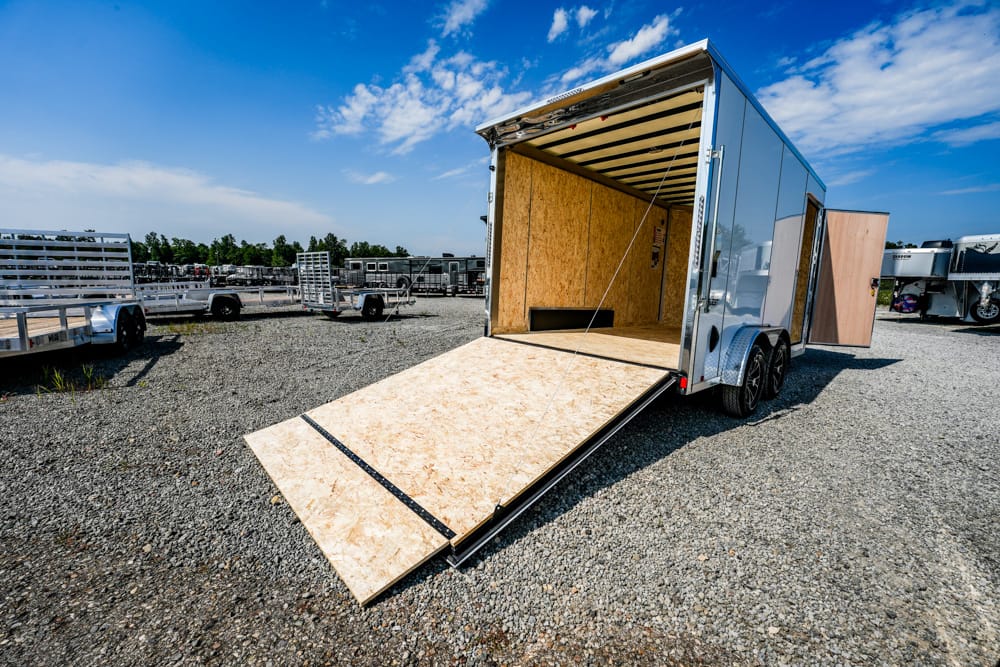 Open aluminum cargo trailer with wooden ramp extended onto a gravel lot under a blue sky.