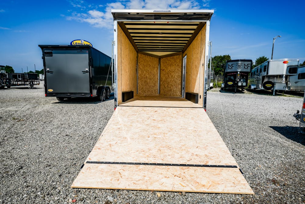 Open empty cargo trailer with plywood interior and ramp extended onto a gravel lot, surrounded by other trailers under a blue sky.
