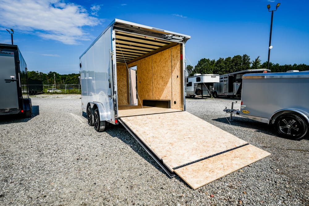 Open white cargo trailer with a plywood interior and lowered ramp on a gravel lot.