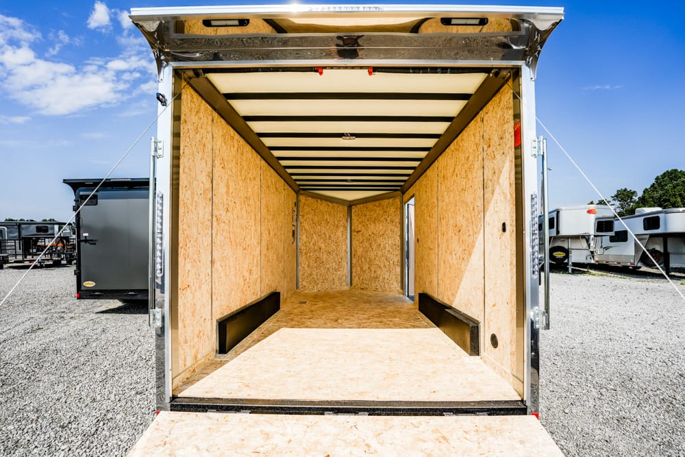 Open empty trailer with wooden walls and ramp, ready for loading in a gravel lot with other trailers nearby.