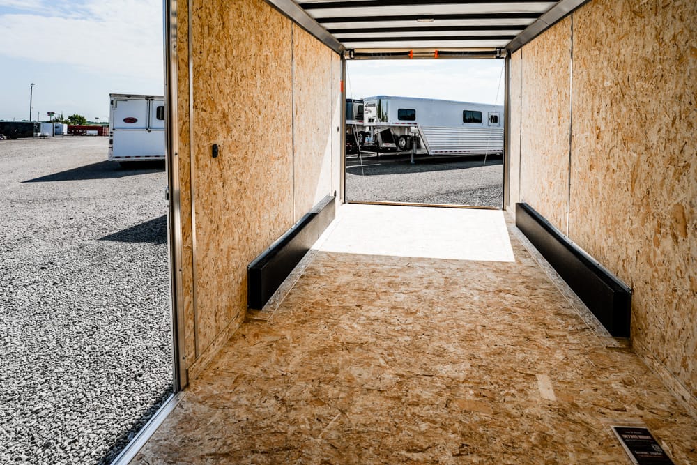 Interior of an empty plywood trailer looking toward an open exit, with a gravel lot and parked trailers visible outside.