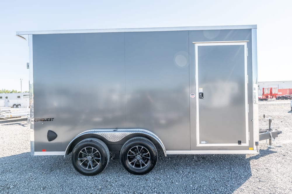Gray enclosed cargo trailer with two axles and a side door on a gravel lot.