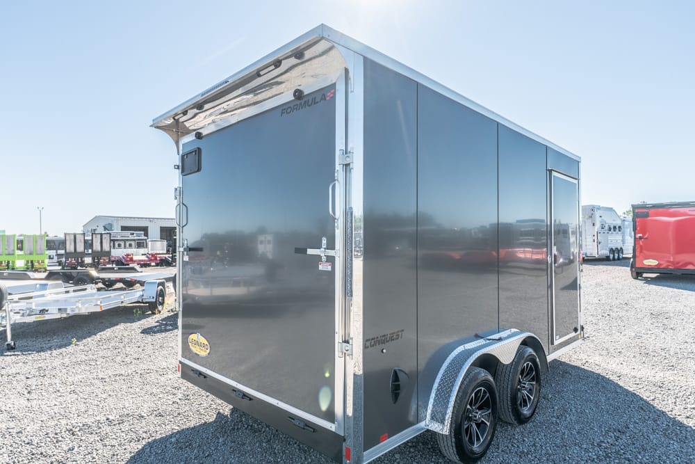 Gray enclosed cargo trailer with dual axles and black alloy wheels on a gravel lot, sunny day in the background, side door visible on the right panel.