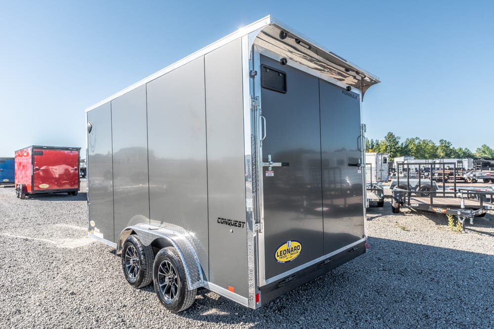 Gray enclosed cargo trailer with tandem wheels parked on a gravel lot, rear doors closed and brand labels visible (Conquest/Leonard).