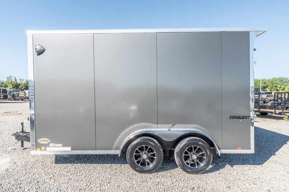 Grey enclosed dual-axle cargo trailer with diamond-plate wheel wells parked on gravel under a clear blue sky.