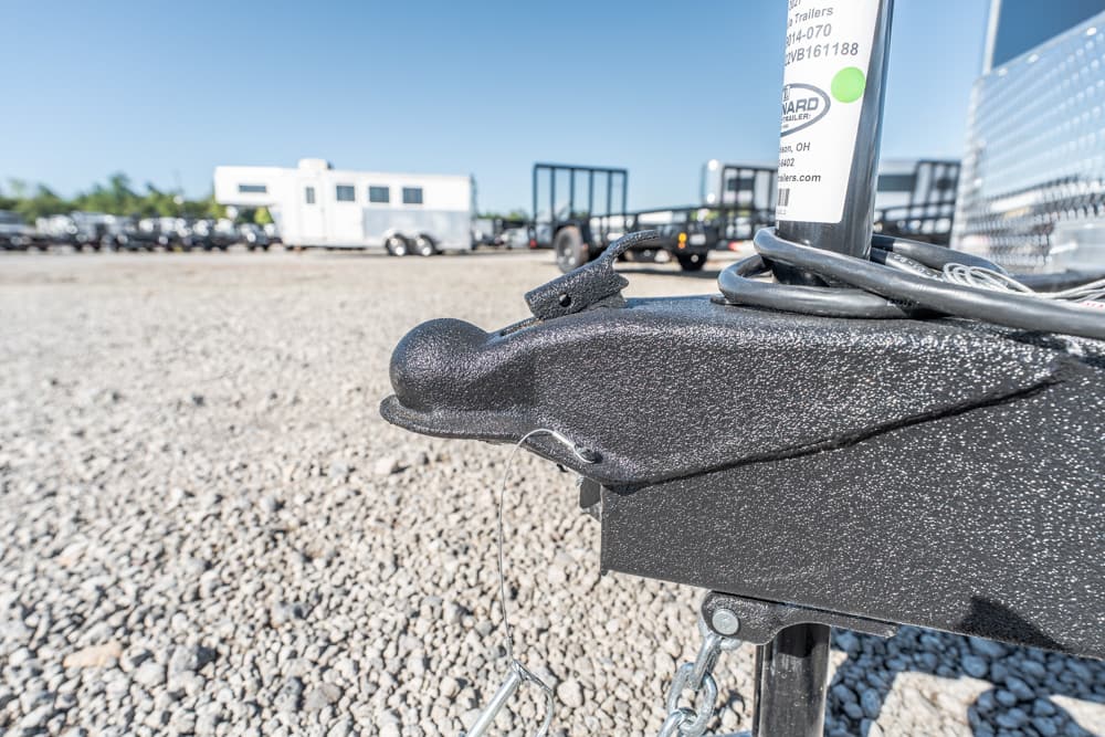 Close-up of a black trailer hitch with safety chains on a gravel lot, trailers visible in the distance.