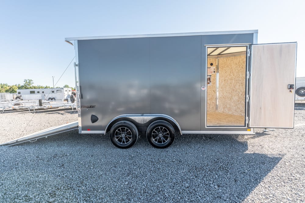 Gray enclosed cargo trailer with dual axles on a gravel lot; side door swing-open reveals plywood interior, ramp extended at left