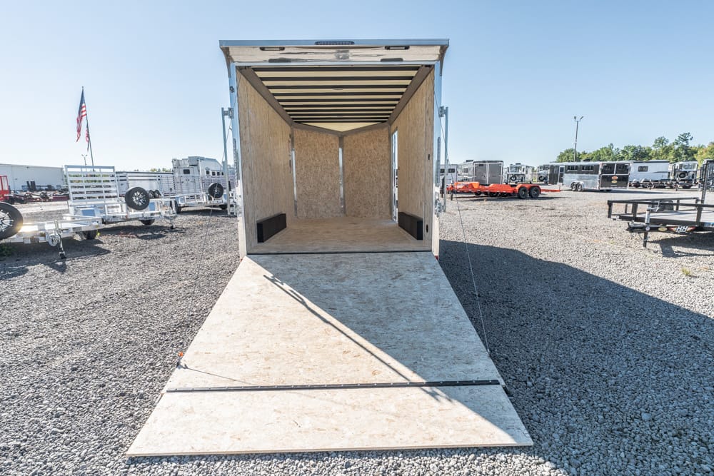 Open cargo trailer with lowered ramp in a gravel trailer yard among other trailers and equipment in the background from a center perspective.