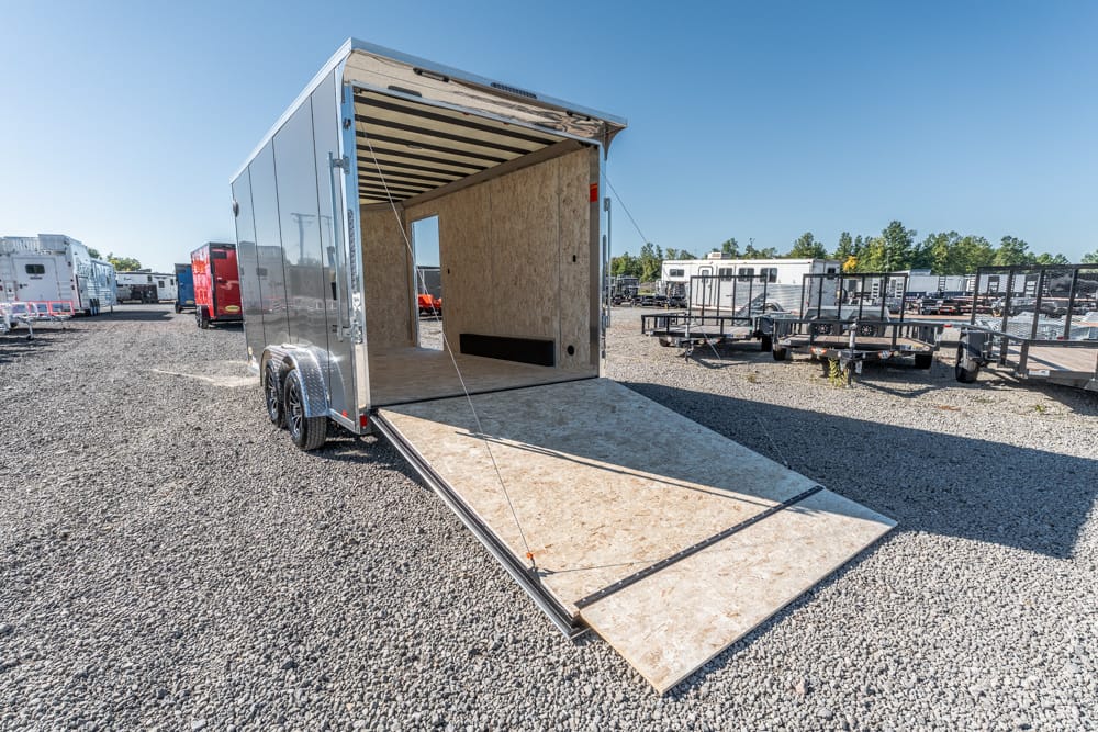 Open silver cargo trailer with its wooden ramp extended for loading on a gravel lot, trailers visible in the background.