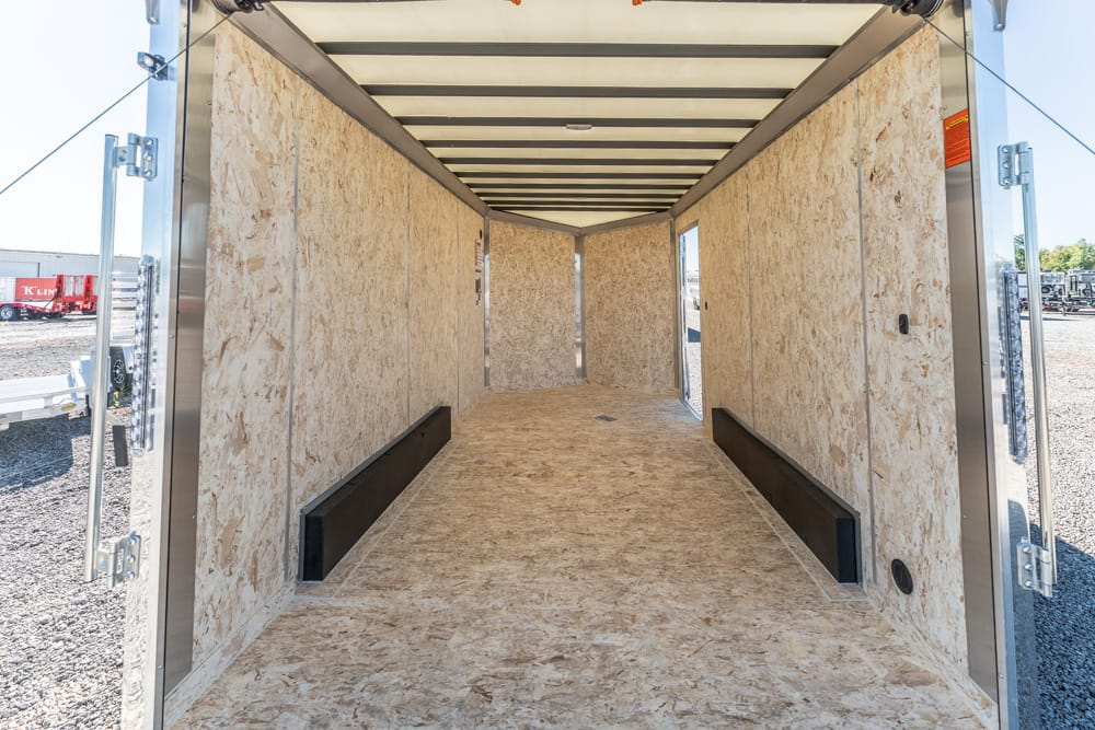 Empty cargo trailer interior with plywood walls and two black wheel wells along the floor, awaiting cargo.