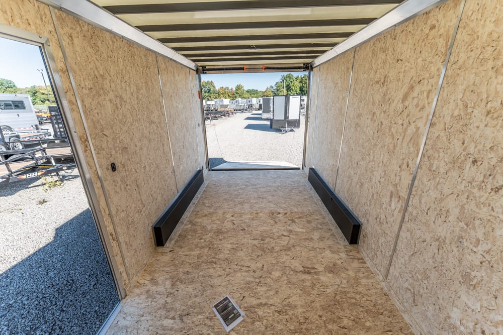 Empty trailer interior with plywood walls, open back to a gravel lot with trailers in the distance; two black wheel wells along the floor.
