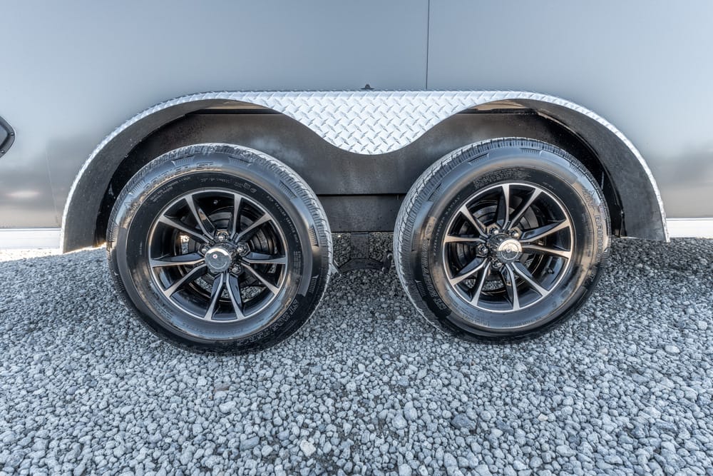 Dual trailer wheels with black rims under a silver trailer and diamond-plate fender on a gravel surface.