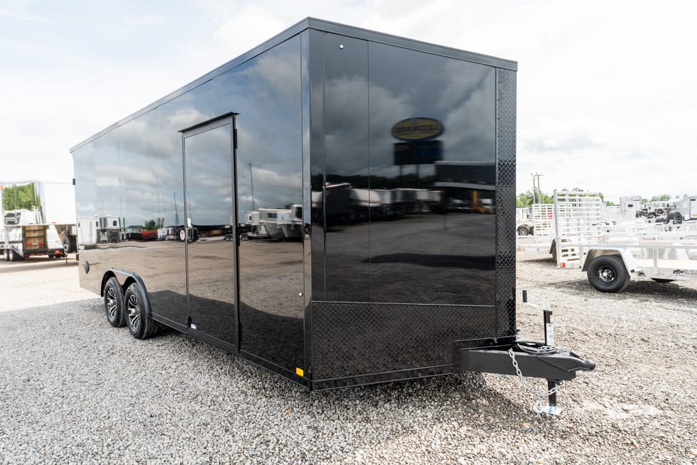 Black enclosed cargo trailer with dual axles parked on a gravel lot; side door, reflective glossy panels, and a front hitch with jack.