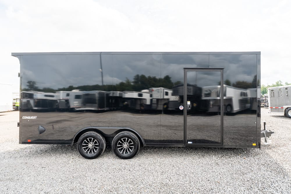 Black enclosed cargo trailer with dual axles and a side entry door, parked on a gravel lot.