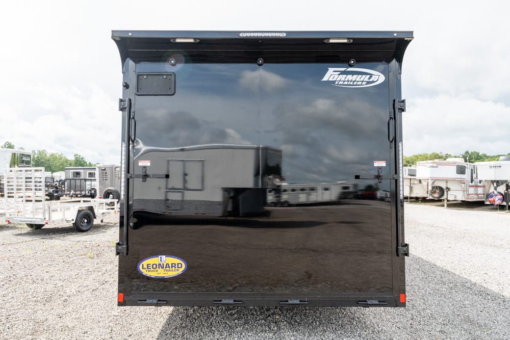Rear view of a black enclosed cargo trailer with Formula Trailers branding and a Leonard sticker, parked on a gravel lot with other trailers nearby.