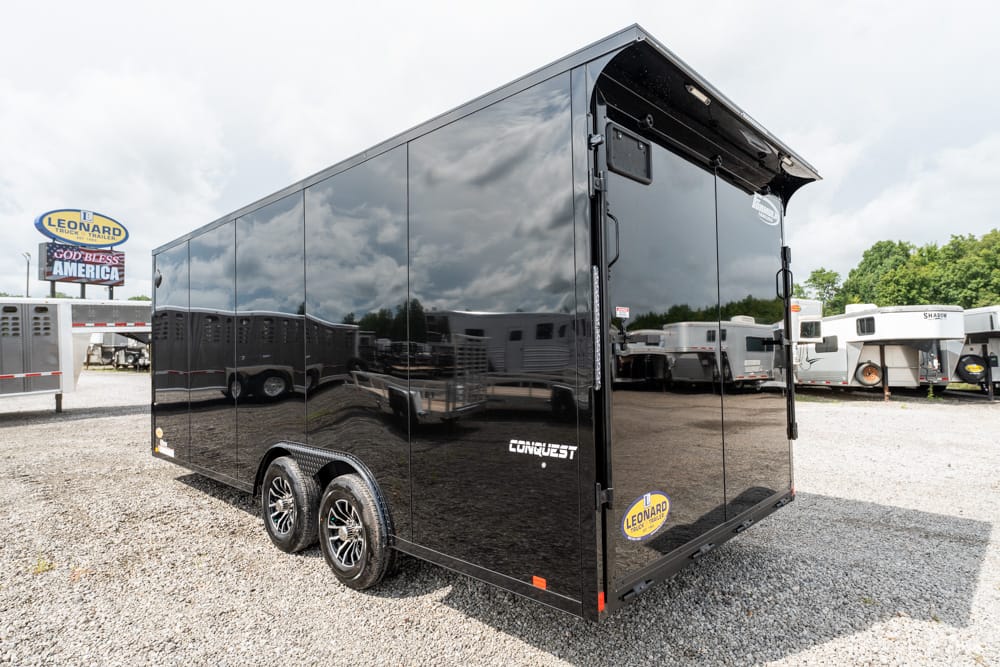 Black glossy enclosed cargo trailer with dual axles on a gravel lot, dealership signs visible in the background.